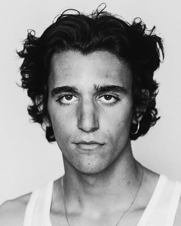 a black and white closeup portrait of a young man with wavy, curly dark hair. he is wearing a simple white tank top and small hoop earrings. his expression is serious, and his gaze is direct. the background is plain and uncluttered, keeping the focus on the subject. the overall aesthetic is clean and somewhat moody.