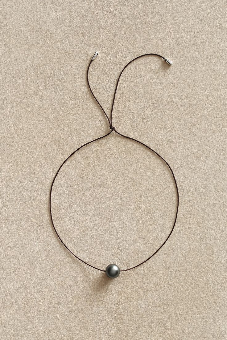 a simple necklace with a single black pearl on a brown cord. the pearl is positioned in the center of the necklace, with the cord tied into a loop at the back of the neck. the necklace is on a light beige background,  giving it a clean and minimalist look.