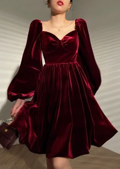 a young woman in a deep red velvet dress with puffed sleeves, a sweetheart neckline, and a midi length poses in a studio. the dress is fitted and flows nicely around her legs. she has her hands on her hips and looks to the side with a slightly posed expression.  the red velvet is soft and shiny and gives the dress a luxurious look.