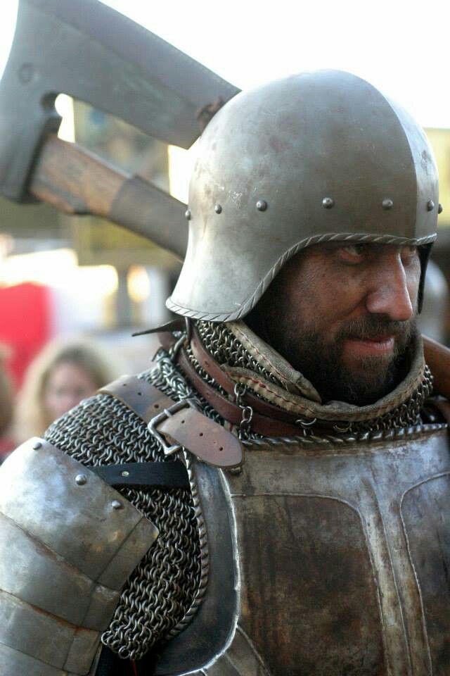a closeup image features a knight wearing full armor, including a helmet and chainmail. the knight has a beard and is holding an axe.
