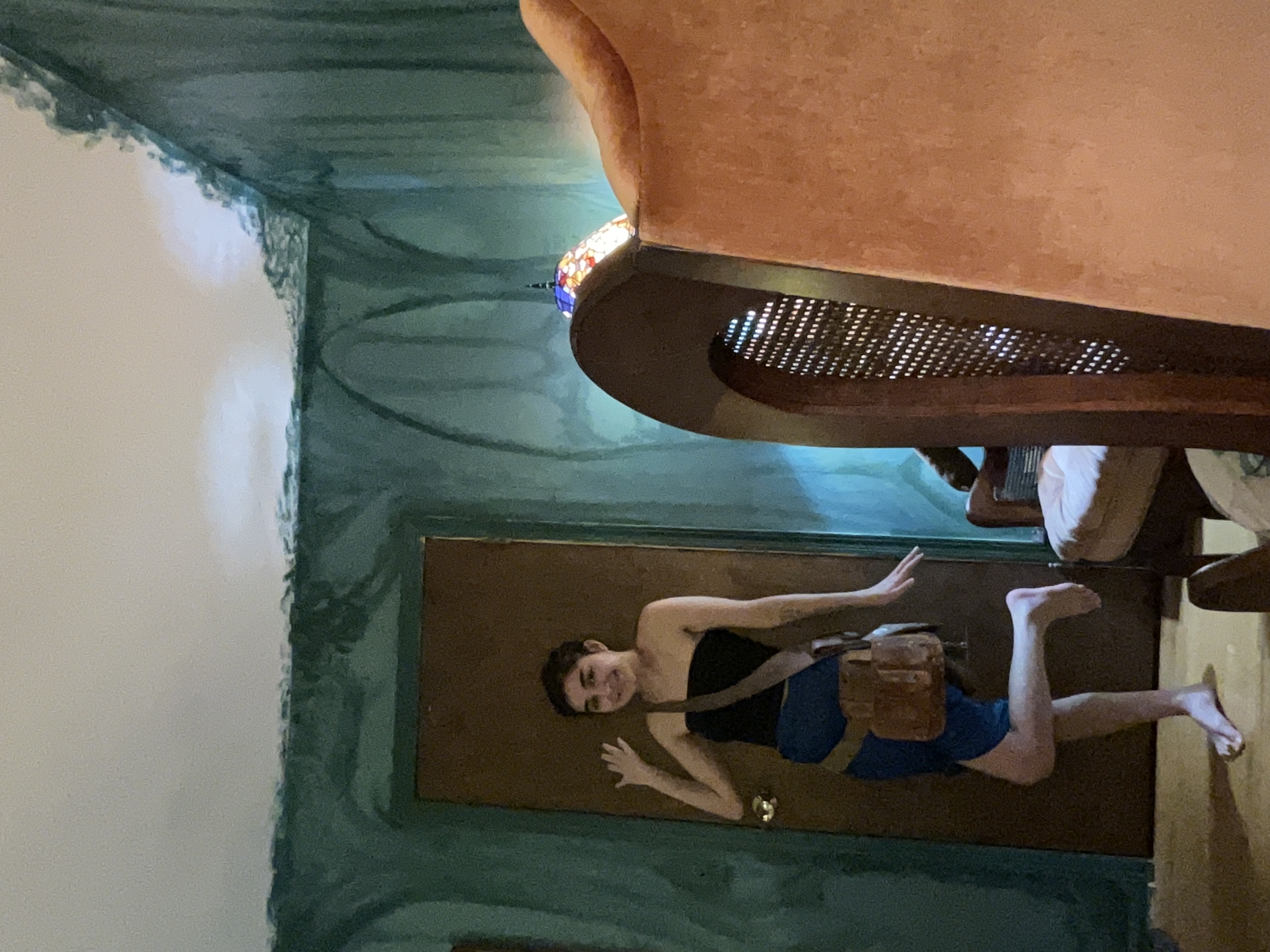 a young woman with short brown hair is standing in a bedroom with uniquely painted walls. the walls feature a dark teal or green color with a swirling, almost art nouveau or art decoinspired design. she is wearing a short dark blue skirt, a black strapless top, and a brown messenger bag. she's barefoot and has one leg bent at the knee.  in the background is a large, orange upholstered armchair with a wooden frame.  the overall ambiance is whimsical and slightly vintage.