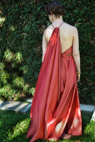 a woman is standing with her back to the camera, wearing a long, draping red dress. the dress has a halter style top and is backless. her hair is styled in an updo. she is standing outdoors in front of green foliage.