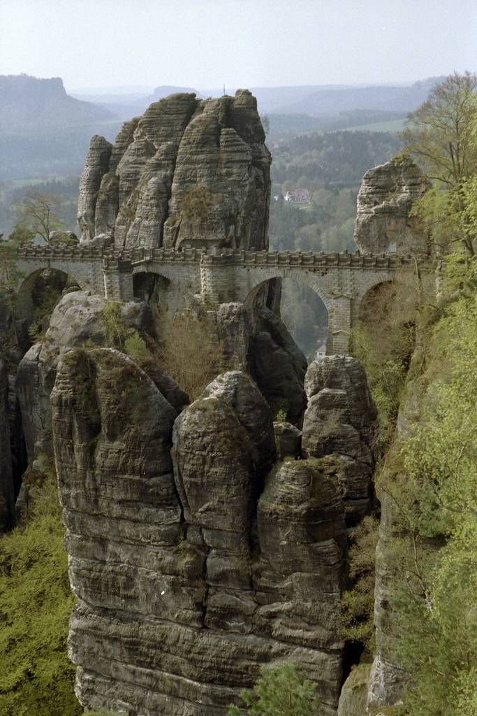the image shows the bastai bridge in germany. the bridge is made of stone and spans across sandstone cliffs. there are trees and nature visible in the image, along with a view of the landscape. the bridge is a popular tourist destination and a historic landmark.
