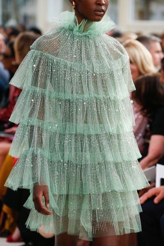 a model on a runway wearing a stunning green tulle dress adorned with sequins. the dress is layered and frilly, creating a dramatic and elegant look.