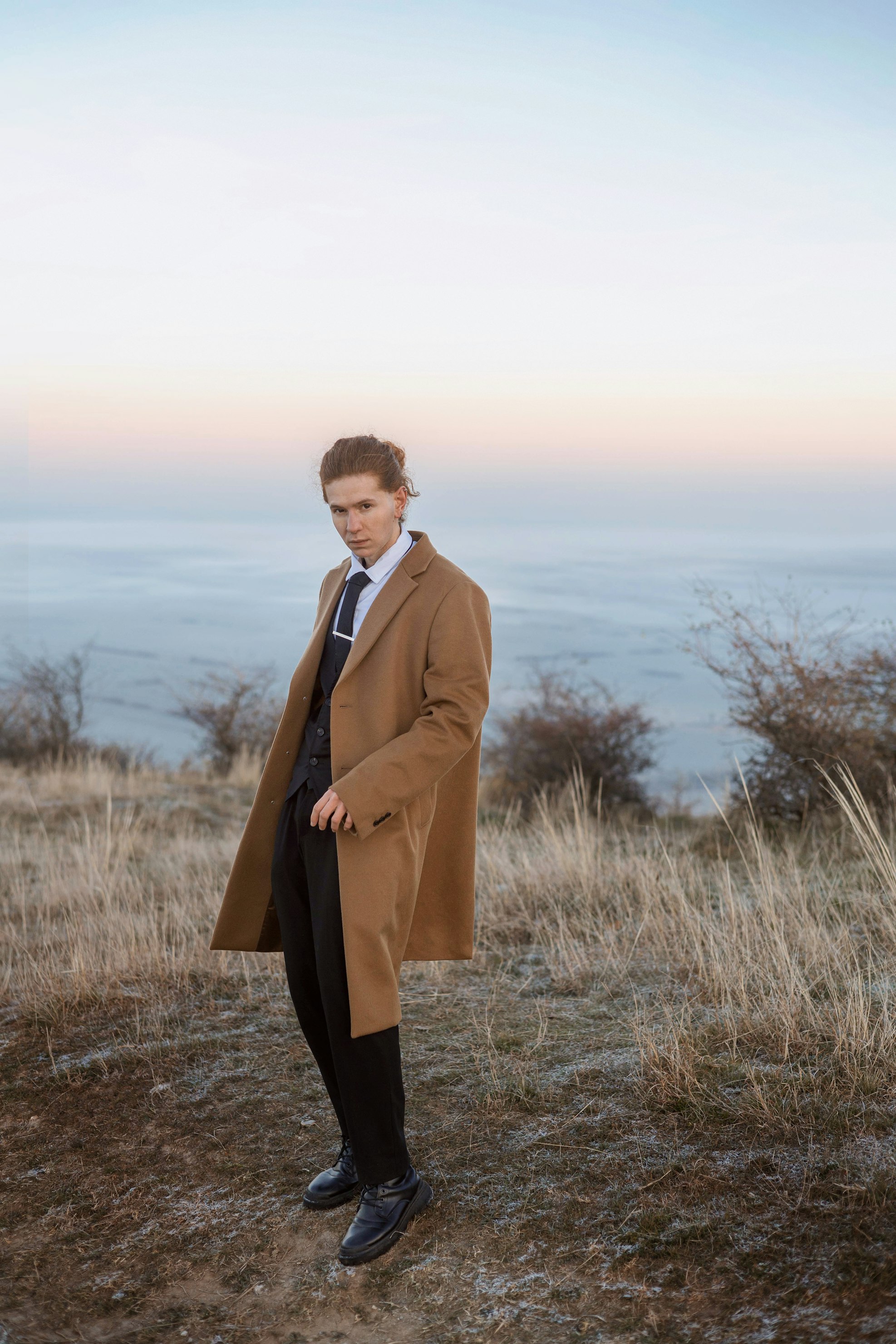 a man in a suit and coat stands in a field. he is wearing a dark vest, white shirt, dark tie, black pants, and a camelcolored coat. the background features a landscape with grass, trees, and a body of water in the distance under a pastel sky.
