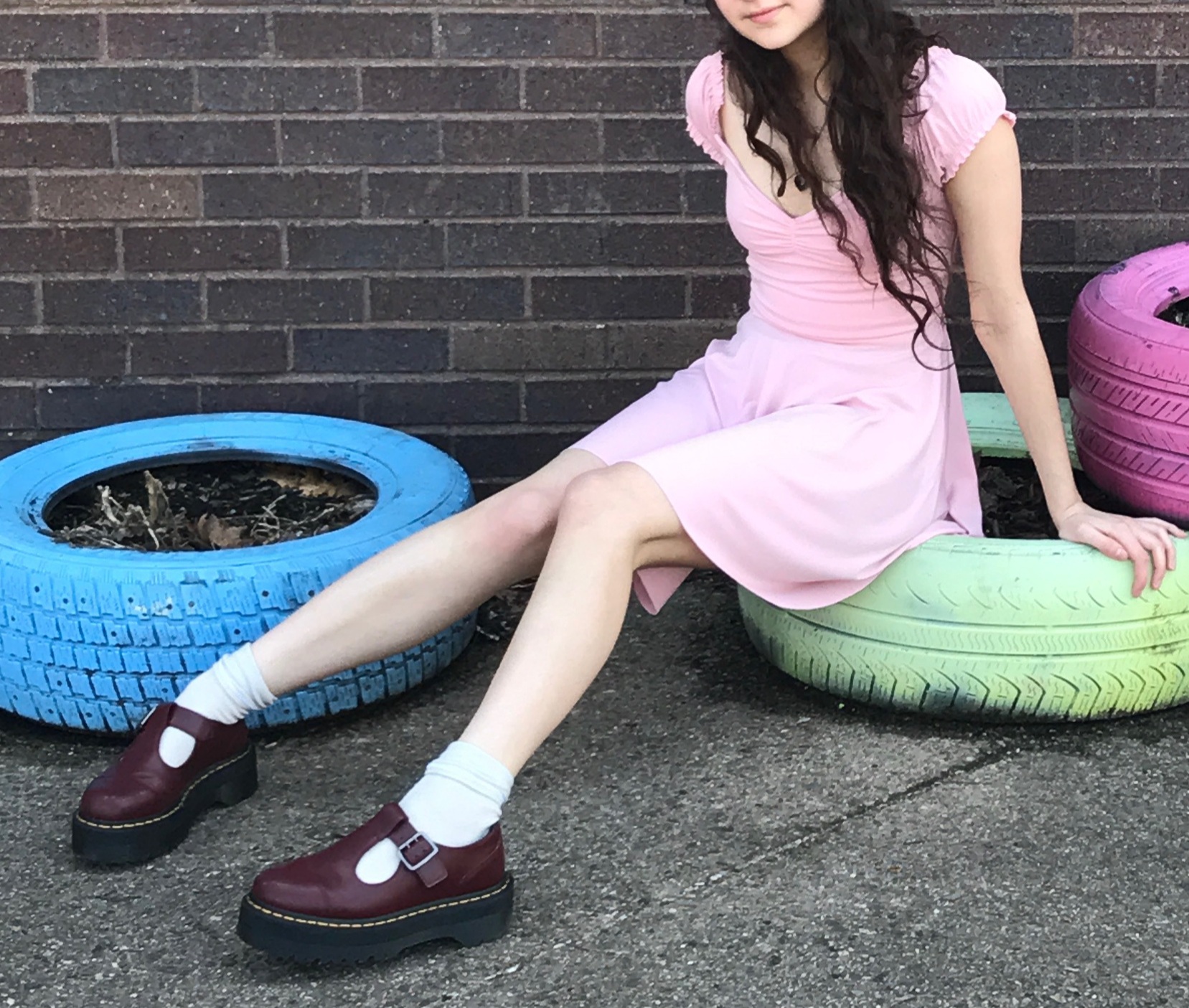 a young woman wearing a light pink dress and dark burgundy dr. martens mary jane shoes sits on repurposed tires used as planters against a brick wall.  the tires are painted different pastel colors. the overall aesthetic is cute and slightly retro.