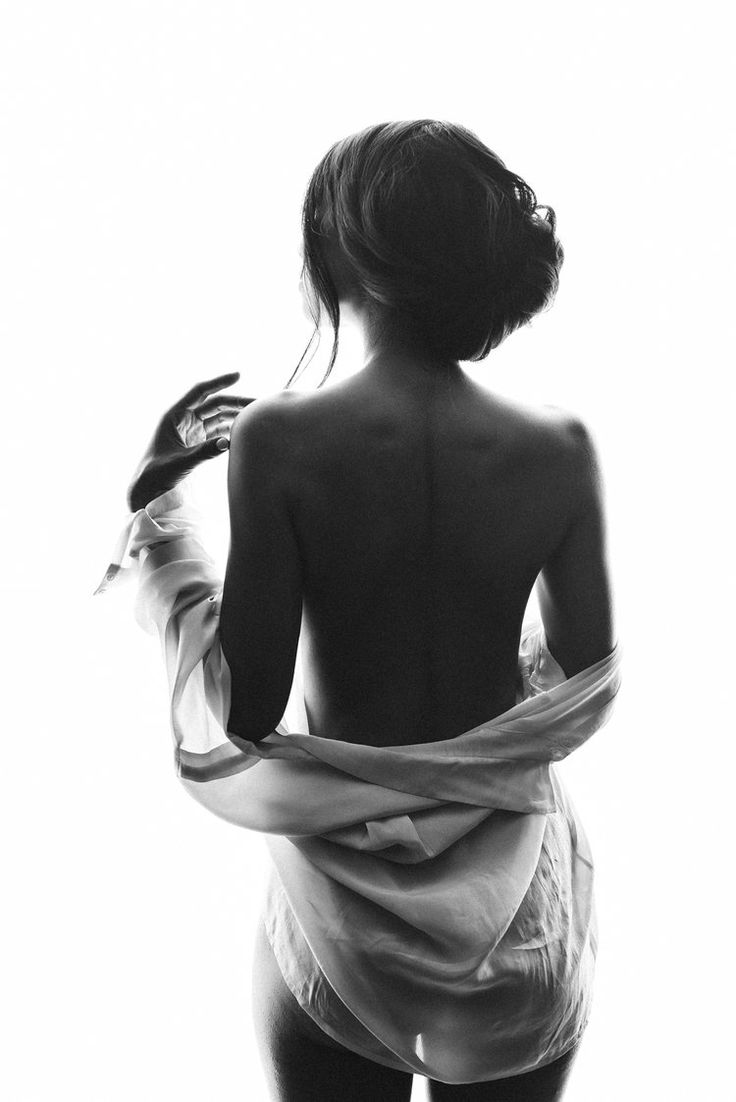 a black and white image of a woman's back. she is nude, but a sheer shirt is draped around her. her hair is styled up, and her hands are gently resting on her shoulders. the background is white, which highlights the contrast of the woman's body. the image is artistic and sensual, highlighting the beauty of the female form.