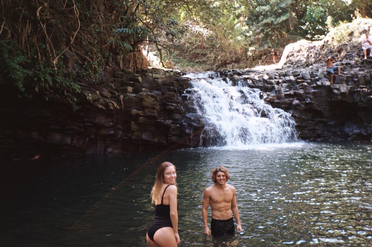 a woman in a black swimsuit and a shirtless man stand in the water in front of a waterfall. there are rocks and trees in the background, and other people can be seen around the waterfall.
