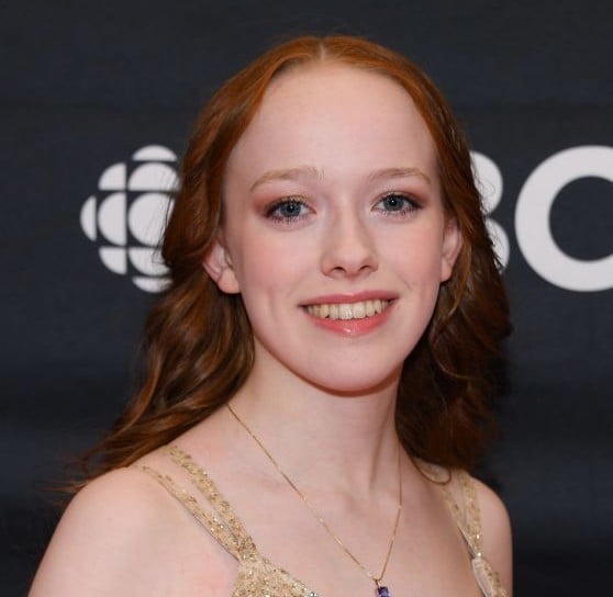closeup portrait of a young woman with long, wavy red hair. she's smiling and wearing a gold dress and a delicate necklace. the background is dark with a cbc logo visible. she looks elegant and happy, possibly at an awards ceremony or gala event.  her makeup is subtle and enhances her natural beauty.