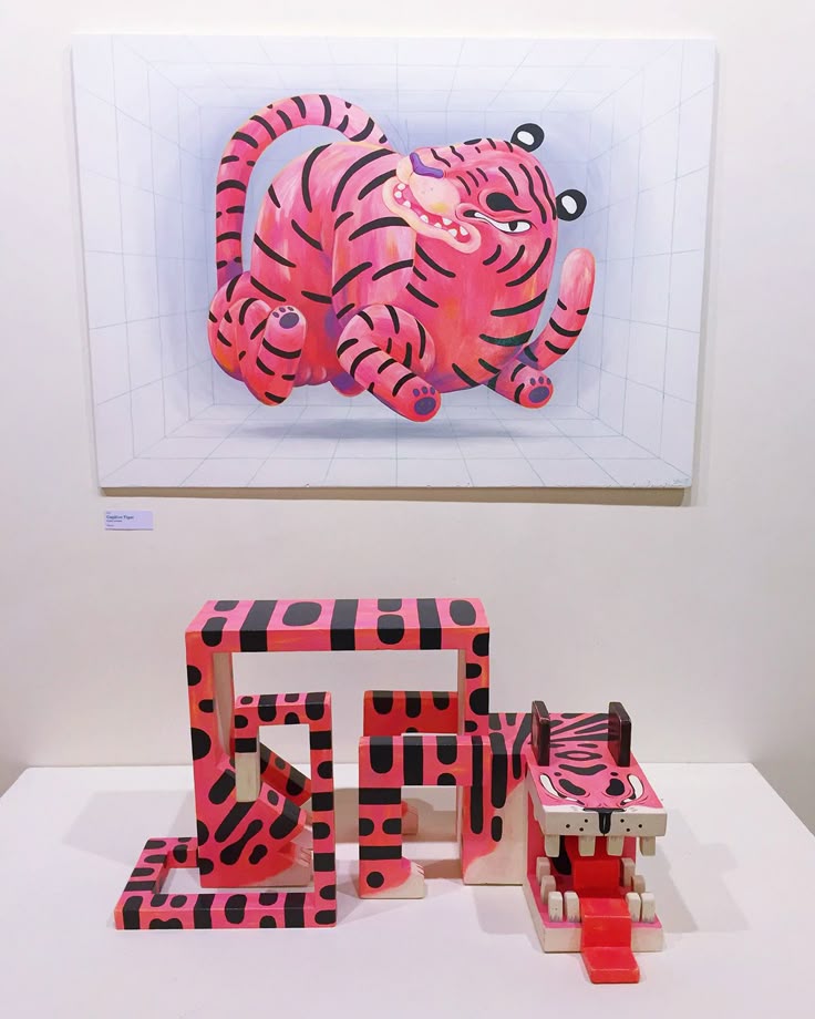 the image shows an art installation in a gallery or exhibition space. on the wall is a painting of a pink cartoonish tiger with black stripes, set within a perspective box. below the painting, on a white platform, there's a wooden sculpture, also pink with black stripes, consisting of abstract geometric shapes and a tiger head with an open mouth.