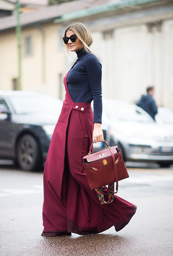 a woman is shown walking outside. she is wearing sunglasses, a black longsleeved turtleneck, and a maroon maxi dress. she is holding a maroon handbag. two cars are parked behind her, and another person is visible in the background.