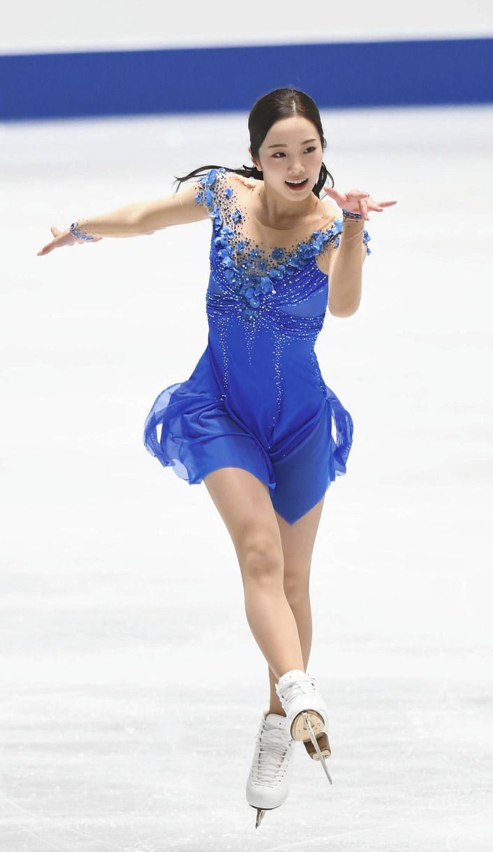 a female figure skater is in midair during her routine. she is wearing a beautiful blue dress with flower detailing. she is smiling as she skates.  the photo was taken at an indoor rink.
