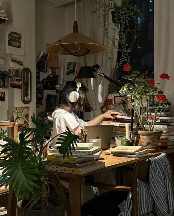 the image shows a young woman studying at her desk in a cozy and welllit room. the desk is cluttered with books, papers, and a laptop. there are plants and framed pictures on the walls, creating a personalized and inviting atmosphere. the woman is wearing headphones, suggesting she is focusing on her work. the warm lighting and abundance of books create a sense of studiousness and comfort.