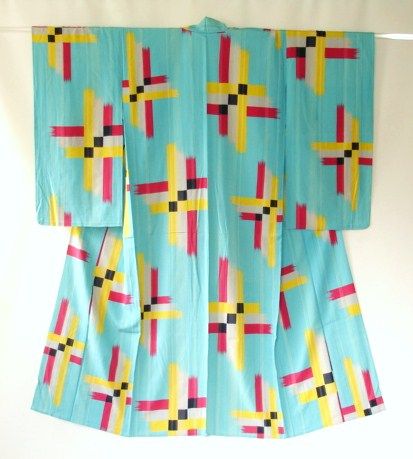 a blue kimono with a repeating geometric pattern of crosses in yellow, red, and black, suspended against a white background.