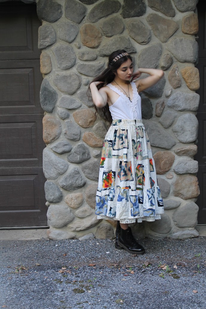 a young woman stands against a stone wall, showcasing her unique fashion sense.  she's wearing a white crocheted top, a long, multicolored patchwork maxi skirt, and black doc martens. a delicate crown or headband adorns her hair. the scene is outdoors, providing a natural backdrop to the model's bohemianinspired attire. the image is styled to evoke a vintage or retro feel.
