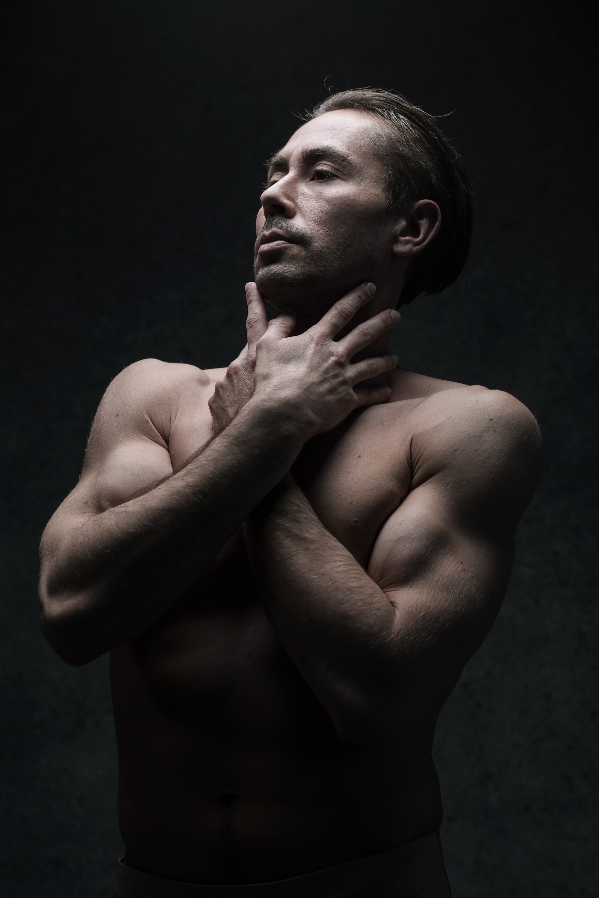 the image showcases a shirtless man standing against a dark, textured background. his pose is contemplative, with his hands gently cradling his neck and his head tilted back. the lighting is dramatic, illuminating his muscular physique and highlighting the contours of his body. the overall mood is both powerful and intimate.