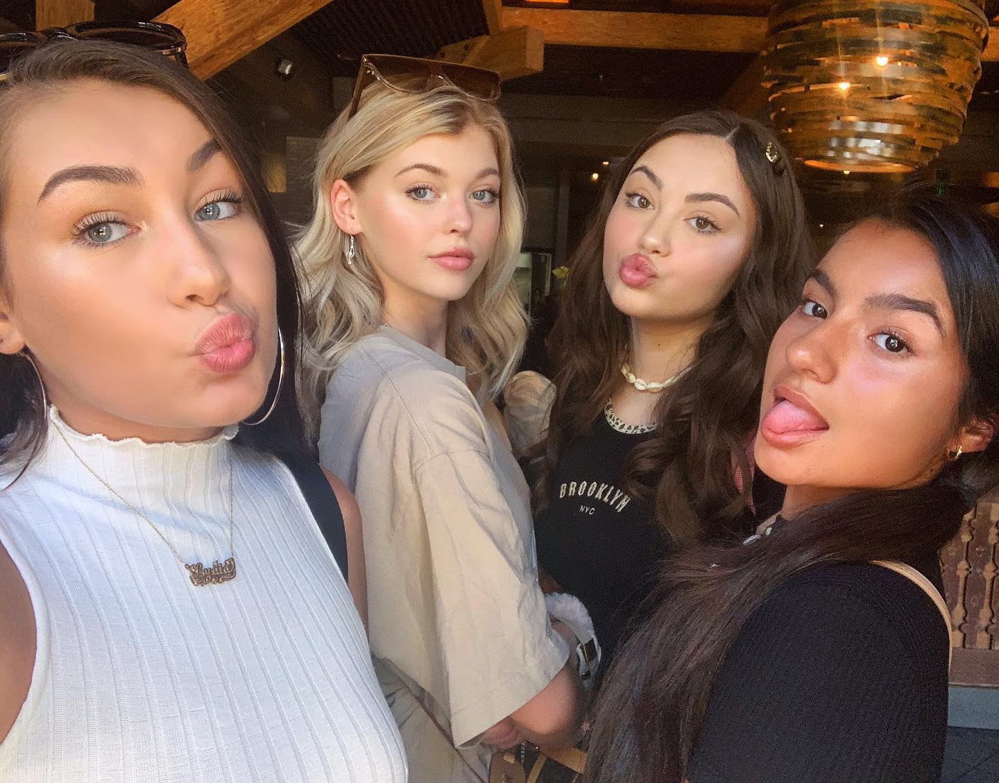 a selfie of four young women posing indoors. the woman on the left is wearing a white top and gold name necklace, pouting with her lips. next to her is a woman with blonde wavy hair, wearing a tan top and small hoop earrings. the third woman is wearing a black top with "brooklyn nyc" graphic, a white seashell necklace, and has a gold hair clip. she is also pouting. the woman on the right has her tongue sticking out and is wearing a black top. they appear to be in a restaurant with a unique light fixture visible in the background.