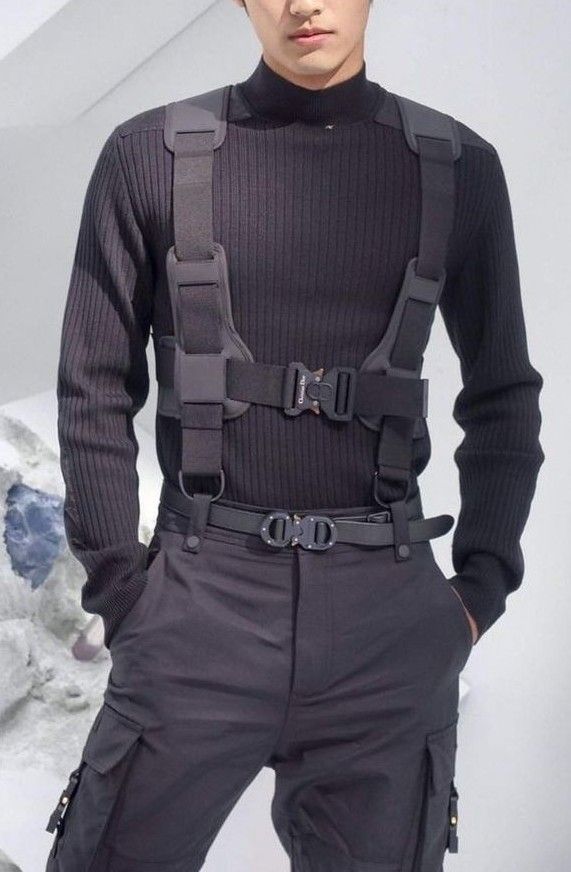 a man in a black turtleneck sweater, black cargo pants with a belt, and a black harness is posing. he is looking at the camera. the man's hair is parted on the side. the background is white.