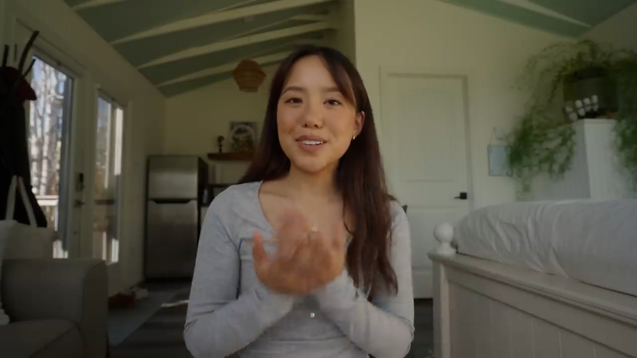 a young asian woman sits in a brightly lit, minimalist bedroom, possibly a tiny house, and claps her hands together while smiling brightly at the camera. her expression and body language convey happiness and excitement, suggesting she may be about to share good news or an announcement.