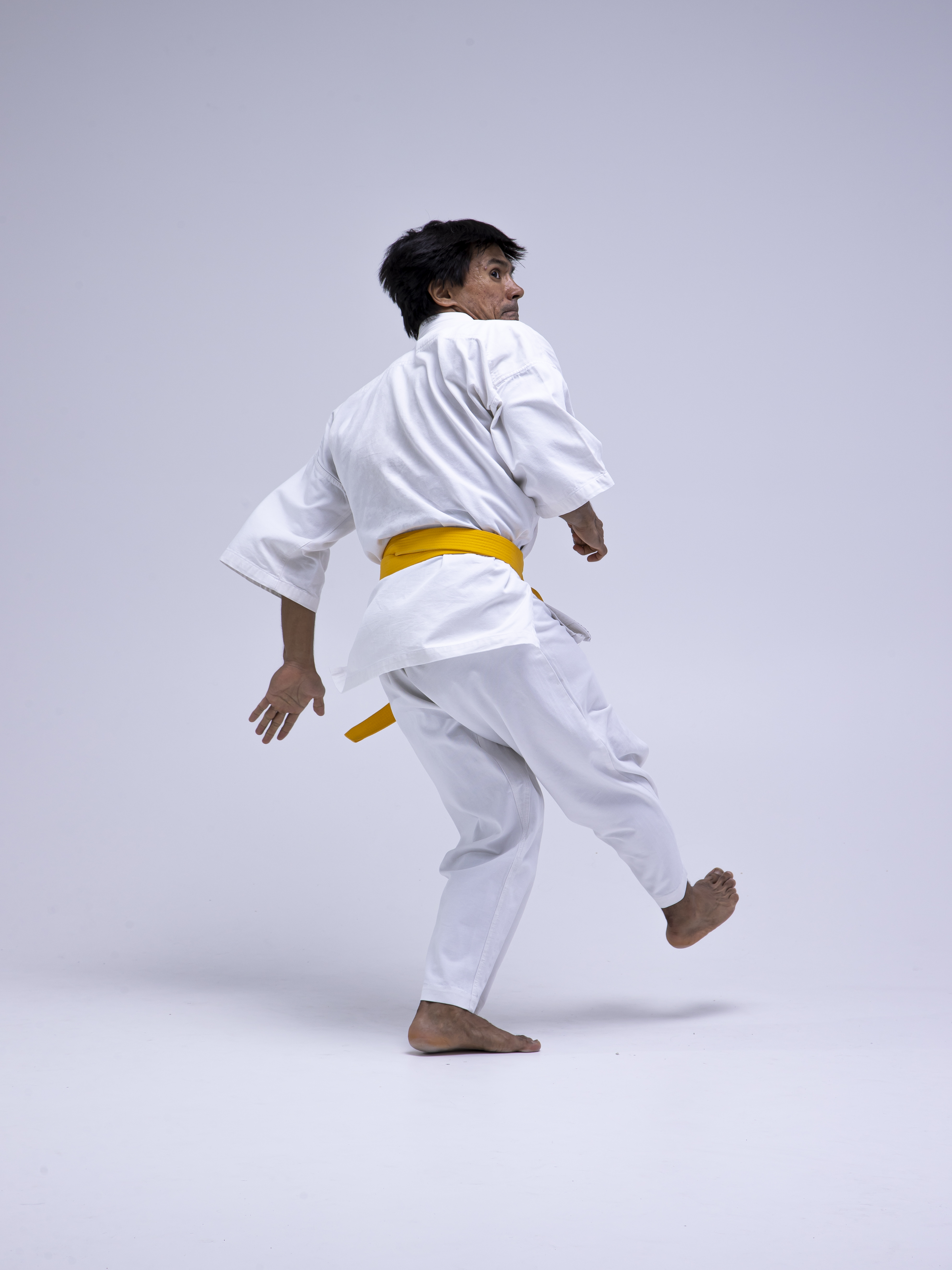 a man in a white karate gi with a yellow belt is performing a kicking technique in a studio. he is barefoot and looking over his shoulder with a focused expression. the background is white. he is in motion, demonstrating discipline, strength, and power.