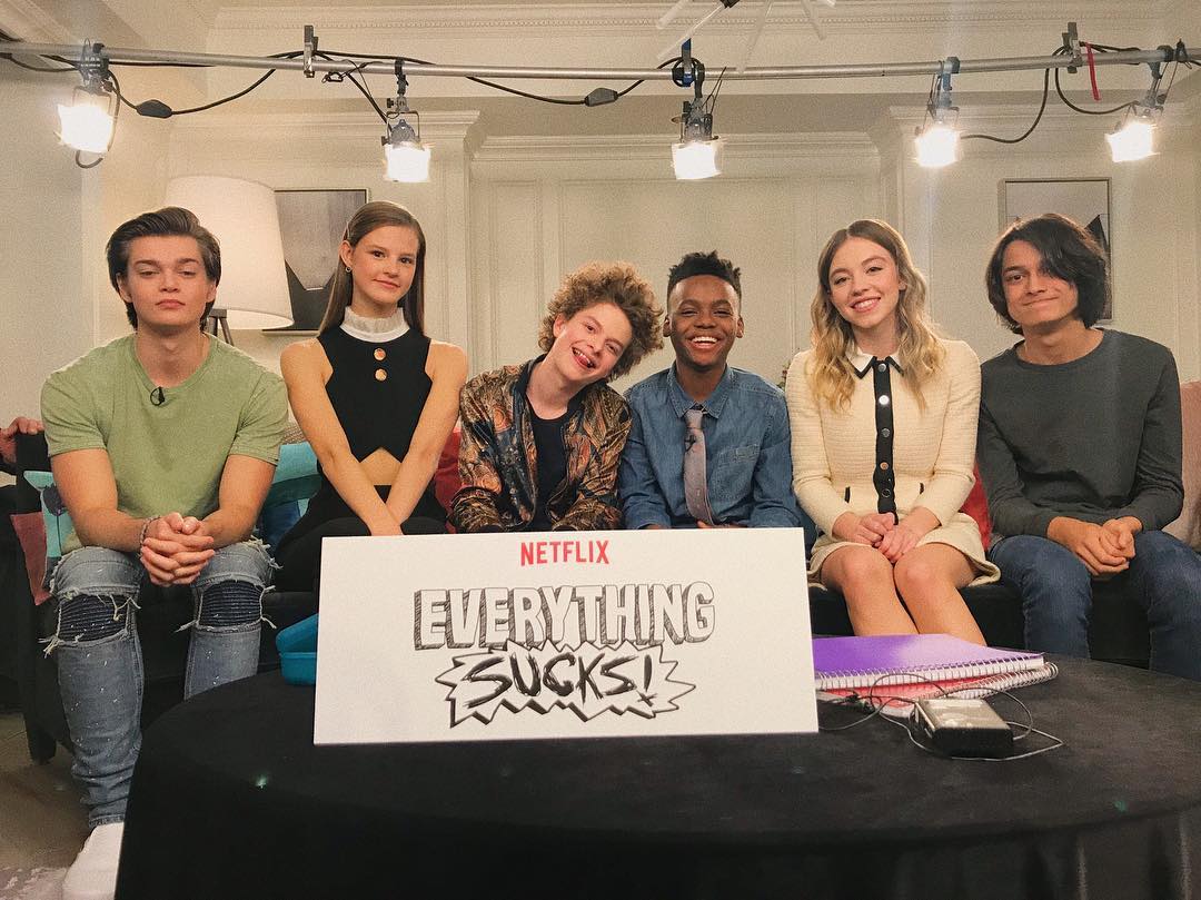 this is a behindthescenes photo of the main cast of the netflix show "everything sucks!"  the actors are posed together on a couch in a room with professional lighting equipment set up. they are holding a whiteboard with the show's title written on it. the overall setting suggests it's a promotional photoshoot or interview setting for the show.  the actors appear relaxed and friendly.
