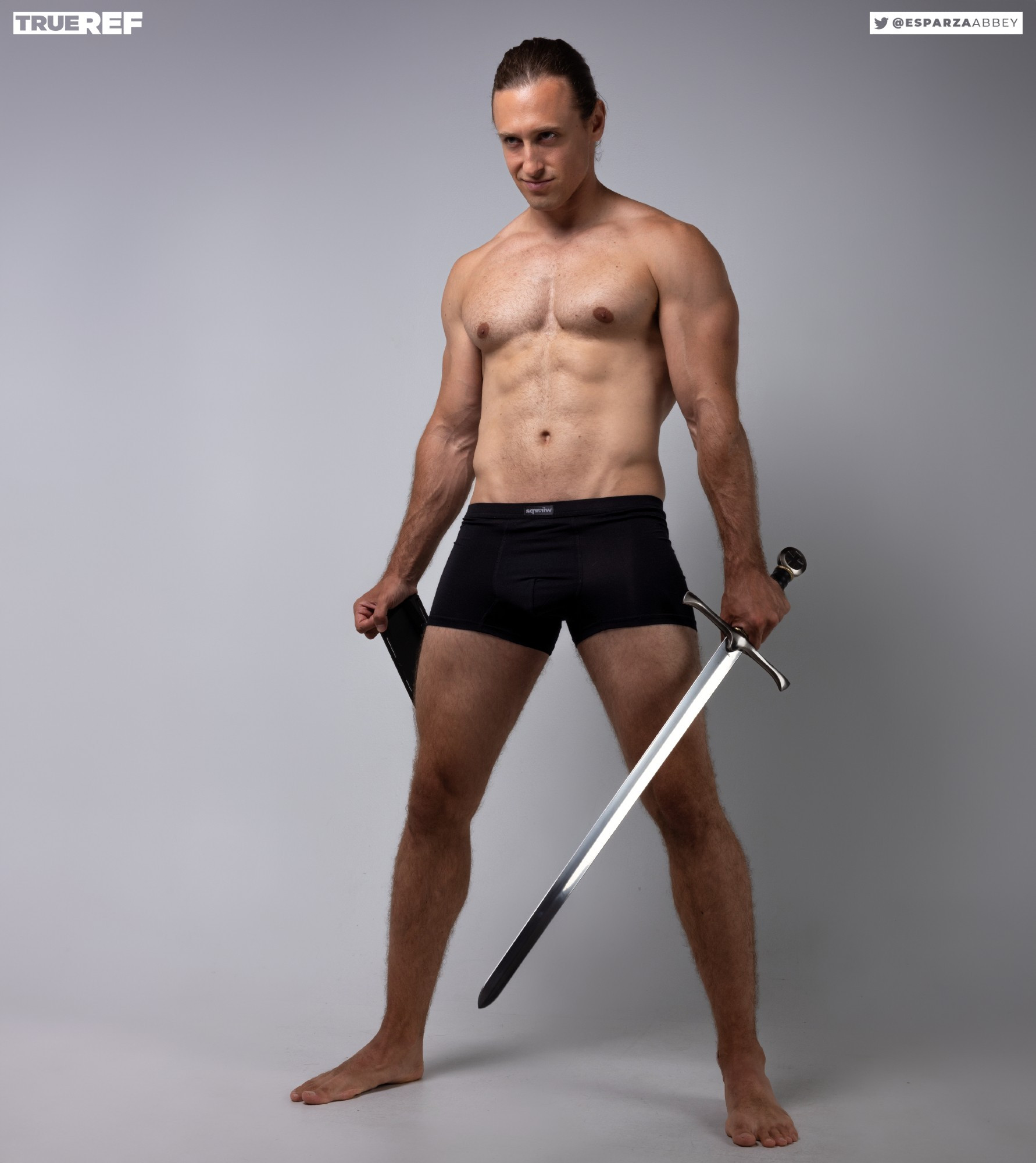 a full shot of a muscular, shirtless man in black boxer shorts holding a long sword. he is posing against a light grey background. the image is professionally lit and the man's physique is very welldefined.  the photo appears to be a reference image for artists or designers, as indicated by the "trueref" watermark and social media handle in the upper corners.