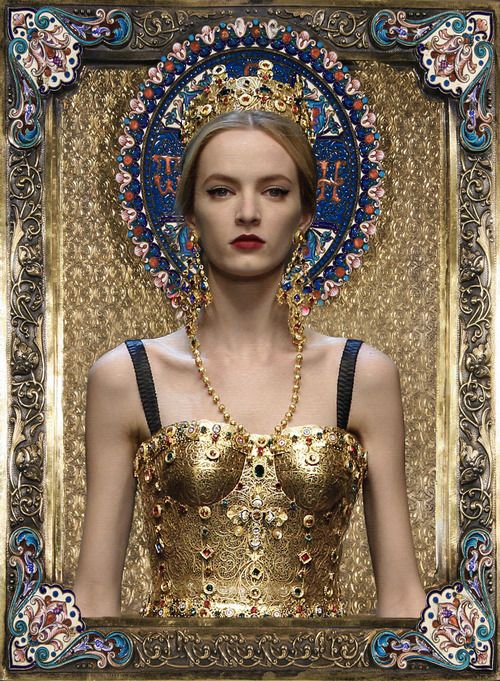 the image showcases a fashion model posed to resemble a religious icon. she wears a lavish gold corset adorned with intricate detailing and precious stones.  a large, ornate crown sits atop her head, framed by a halolike structure that is richly decorated with blue, pink and gold enamel. the entire look is reminiscent of byzantine art. the model is presented within a heavily ornate, painted frame, further enhancing the religious icon effect. the overall aesthetic is one of opulence and high fashion, characteristic of dolce & gabbana's style.