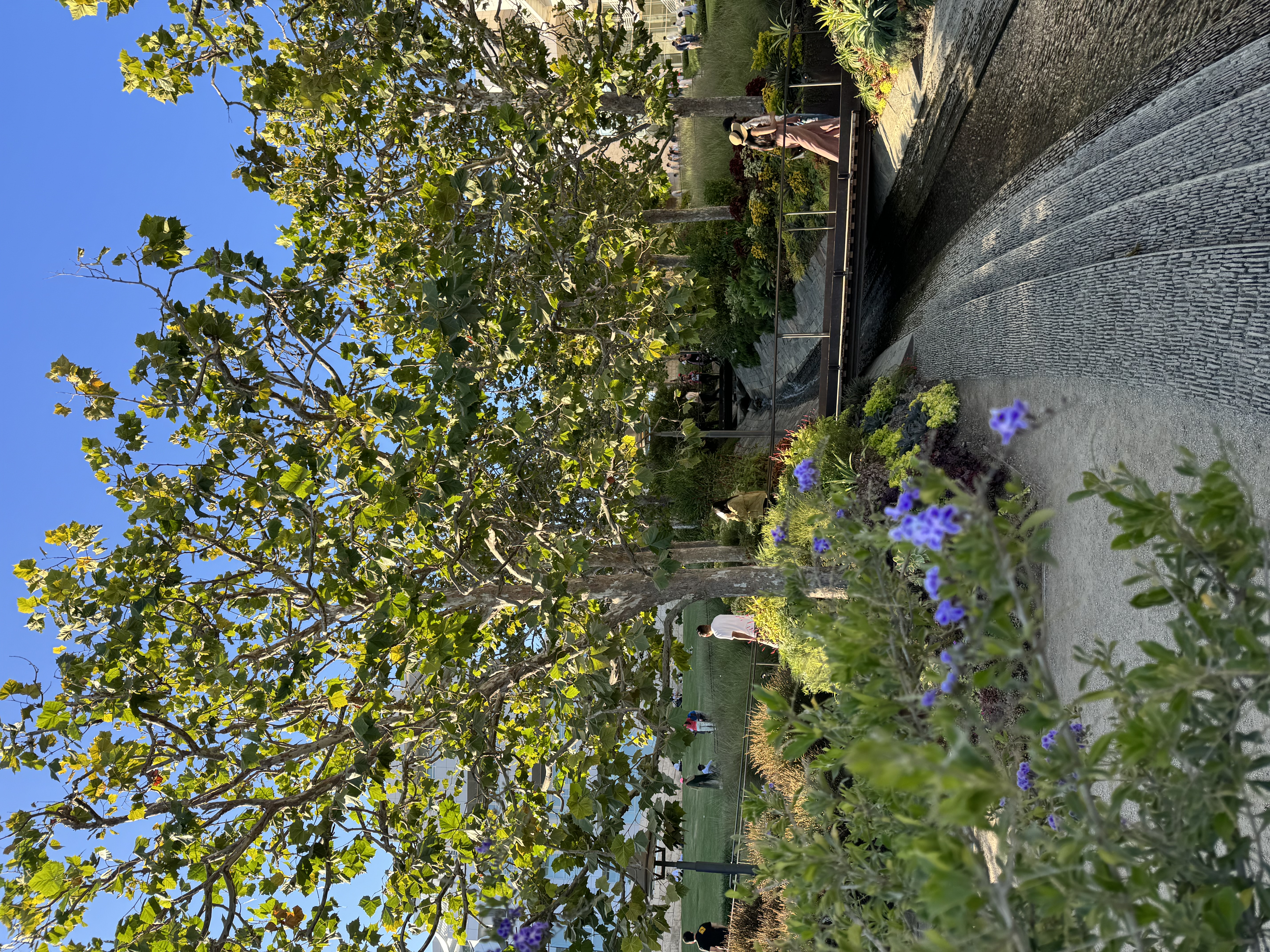 the image showcases a picturesque park scene bathed in warm sunlight. a paved path, flanked by vibrant green grass and colorful flower beds, leads to a wooden bridge. a gentle stream of water flows under the bridge, creating a serene atmosphere. towering trees with lush green foliage provide ample shade and frame the scene. people can be seen strolling along the path, enjoying the pleasant ambiance. the blue sky peeks through the leaves, adding a touch of vibrancy.