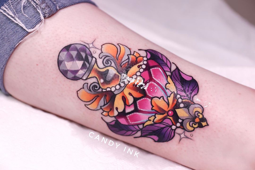 a closeup photograph showcasing a vibrant tattoo of an ornately decorated perfume bottle, shaped like a heart, inked on the inner forearm of a person.  the tattoo is done in a neotraditional style, featuring bold outlines and a bright, colorful palette. the bottle is adorned with a large jewellike stopper and intricate gold and purple detailing.
