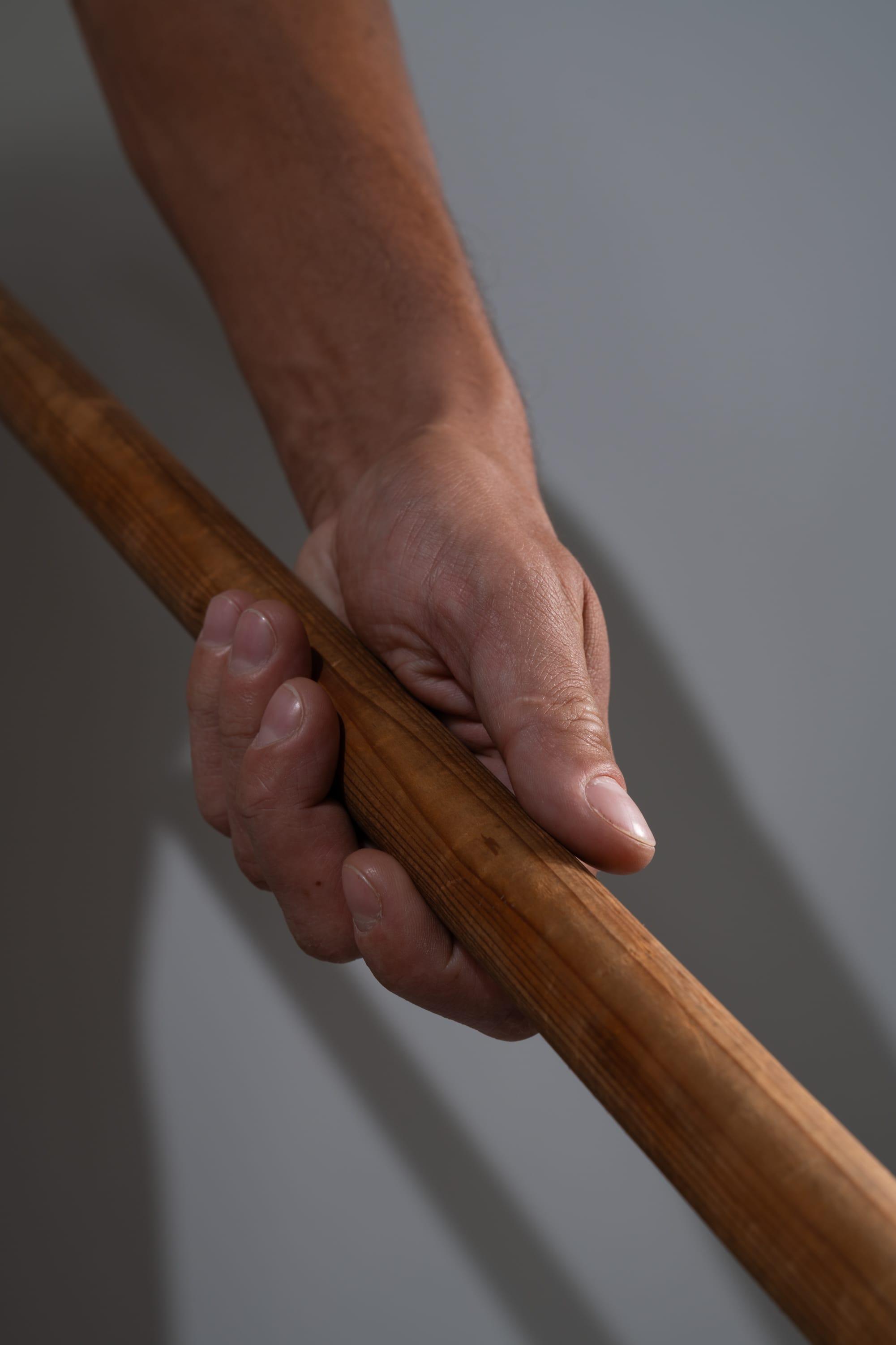 the image shows a closeup view of a man's hand firmly gripping a thick wooden stick. the stick is positioned diagonally across the frame, with the man's hand holding it near the top. the man's arm extends out of the frame, suggesting a sense of strength and control. the background is a neutral gray, emphasizing the focus on the hand and the stick. the studio lighting creates a clear and welldefined image, highlighting the textures of the wood and the skin.