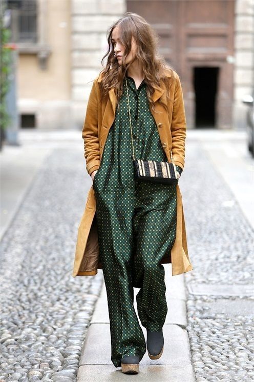 a fullbody shot of a stylish woman walking down a city street. she's wearing a mustardcolored long coat over a dark green patterned jumpsuit. her shoes appear to be clogs or similar footwear. she carries a small, patterned handbag slung across her body. the setting appears to be a european city, with cobblestone streets and oldlooking buildings in the background. the overall style is relaxed yet fashionable, suggestive of autumn or fall weather. the woman's hair is long and wavy, and her expression is somewhat pensive.