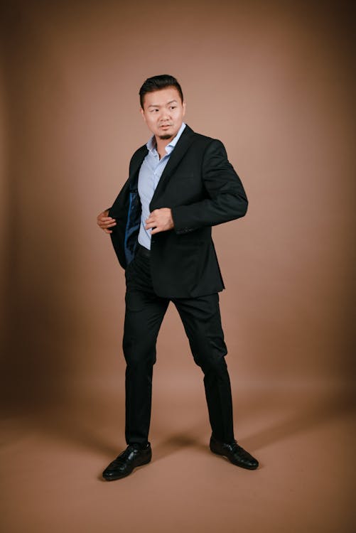 a fullbody studio shot of an asian man in a black suit with a light blue shirt, dress shoes, standing against a plain brown background. he is looking away and opening his suit jacket.