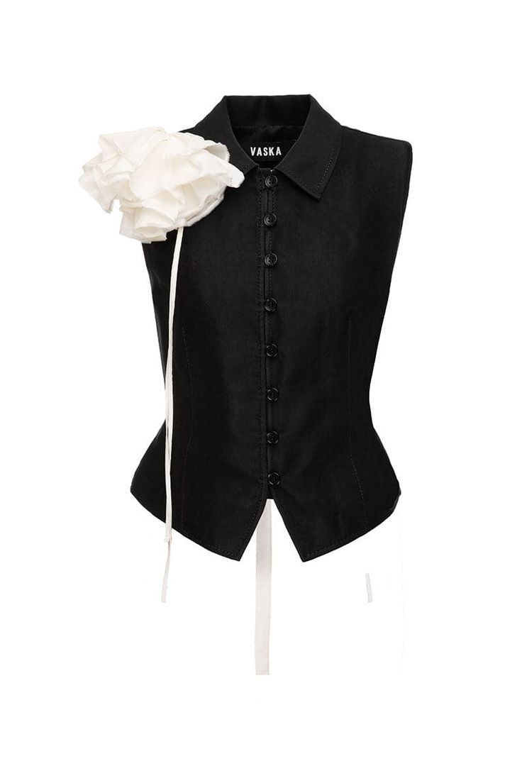 a black sleeveless top with a buttondown front and a large white flower detail on the left shoulder. the brand name vaska is visible on the label near the collar.