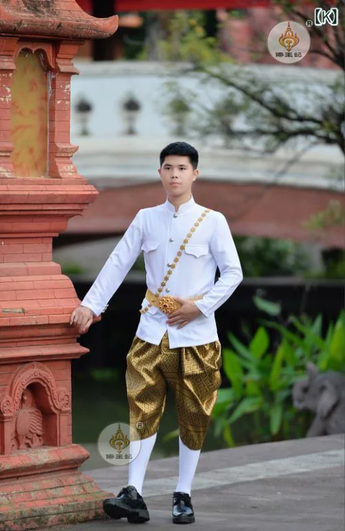 a man is dressed in traditional thai clothing, standing in front of a brick structure. he is wearing a white shirt with gold trim and gold pants. he is looking at the camera.