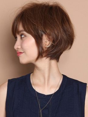 a young woman with short brown hair is looking down. she has a neutral expression on her face. she is wearing a black top and a necklace and earrings. her hair is styled in a bob. she is standing in front of a plain background. the image is a closeup portrait of the woman. she is standing in a studio.