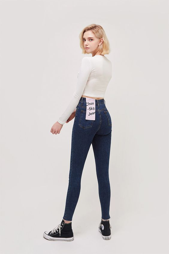 a blonde woman is standing with her back to the camera, showcasing a pair of formfitting skinny jeans. she is wearing a white longsleeve top that ends just above her waist. her jeans have a high waist and are a dark blue wash. she has a small pink tag attached to the back right pocket that says "chuu 5kg jean." she is wearing black and white converse sneakers and is posing against a plain white background.