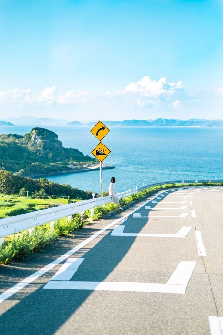 a scenic shot of a winding road next to a coast with a woman sitting on the railing, with signs for a curve and a 10% steep grade. the road has white lines marking the lane and edge. in the background, a vast ocean stretches to the horizon, with mountains and islands visible in the distance. the sky is a mix of blue and white clouds, enhancing the natural beauty of the location.