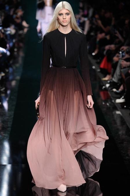 a blonde model walks the runway wearing a long, black and pink ombre gown. the gown has a fitted bodice and a flowing skirt. the model is wearing minimal makeup and her hair is styled in a straight, sleek manner. she is looking straight ahead, with a confident expression on her face. the background is blurred, showcasing the excitement of a runway show.