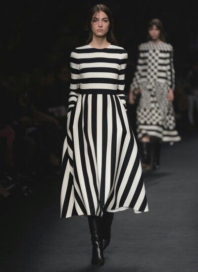 a fashion model walks down the runway in a black and white striped dress and black boots. she is posing for the camera. the background is blurred, but there is another model behind her. the model's hair is long and brown. she is wearing a black and white dress. the dress has horizontal black and white stripes and a wide black stripe around the bottom of the skirt. the dress is tightfitting in the torso and looser in the skirt. the model is wearing black boots with heels.