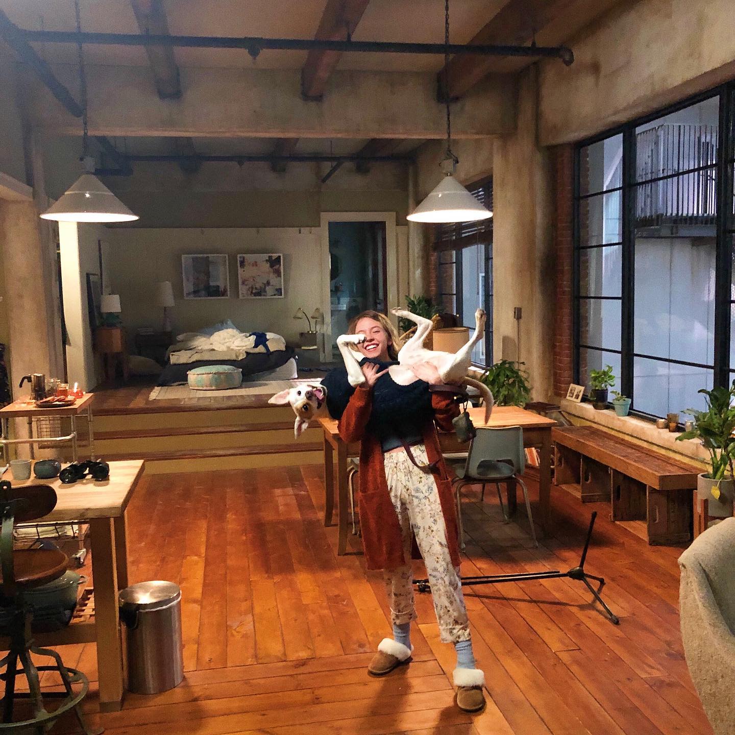 the image shows a happy young woman playfully holding a white dog upside down in a spacious, loftstyle apartment. the woman is smiling broadly and wearing a brown cardigan over floral pajamas and slippers. the apartment features hardwood floors, large windows letting in plenty of natural light, and a rusticindustrial interior design.  the scene appears to be a film or television set, given the design and staging. the overall mood is cozy, comfortable, and lighthearted.