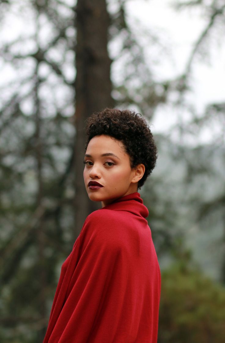a woman with short curly hair and wearing a red turtleneck poncho is standing in a forest, looking back over her shoulder at the viewer. she has a serene expression on her face. the background is blurred, creating a sense of depth.