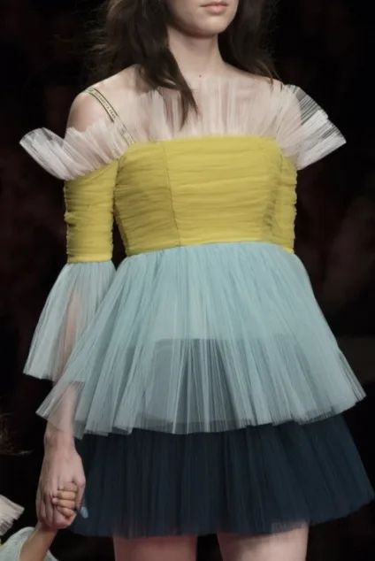 a beautiful woman on the runway wearing a layered, tiered tulle dress with a yellow bodice and a blue and green skirt. the dress is designed with ruffles and a sweetheart neckline.