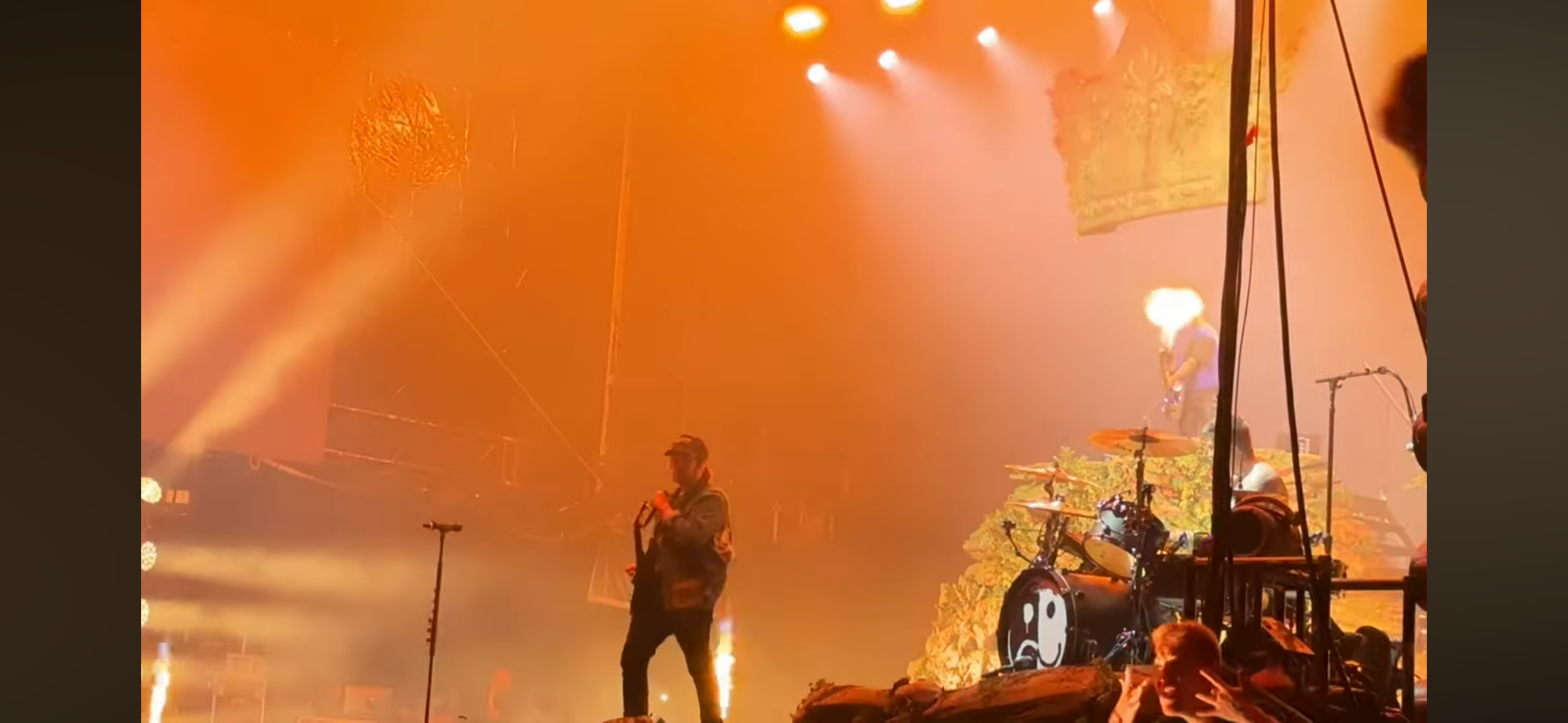 a live music performance with a band on stage. the stage is bathed in orange light and smoke, with pyrotechnics erupting. the lead singer is on the left side of the stage, playing guitar and singing, and the drummer is on the right side. the crowd is visible in the background, cheering on the band.