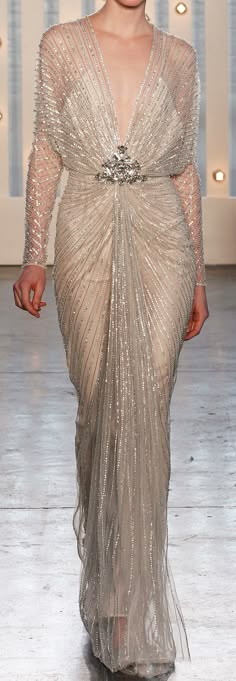 a female model is wearing a luxurious, silver and grey, sequined gown with a deep vneckline.  she is posed with her weight on one leg,  a classic runway pose, highlighting the beauty and texture of the gown. the background is a plain white, highlighting the model and her clothing.  she is ready for a glamorous party or special event.