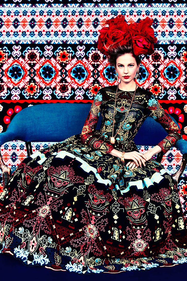 the image is a highfashion photograph of a model wearing an intricately embroidered gown. the dress features a vibrant mix of colors, predominantly red, black, and gold, with intricate geometric and floral patterns reminiscent of mexican or south american folk art. she wears a large, dramatic headpiece of red flowers. the model is seated on a blue sofa against a backdrop of a similarly patterned textile. the overall effect is one of striking visual opulence and artistic flair, typical of editorial fashion photography.