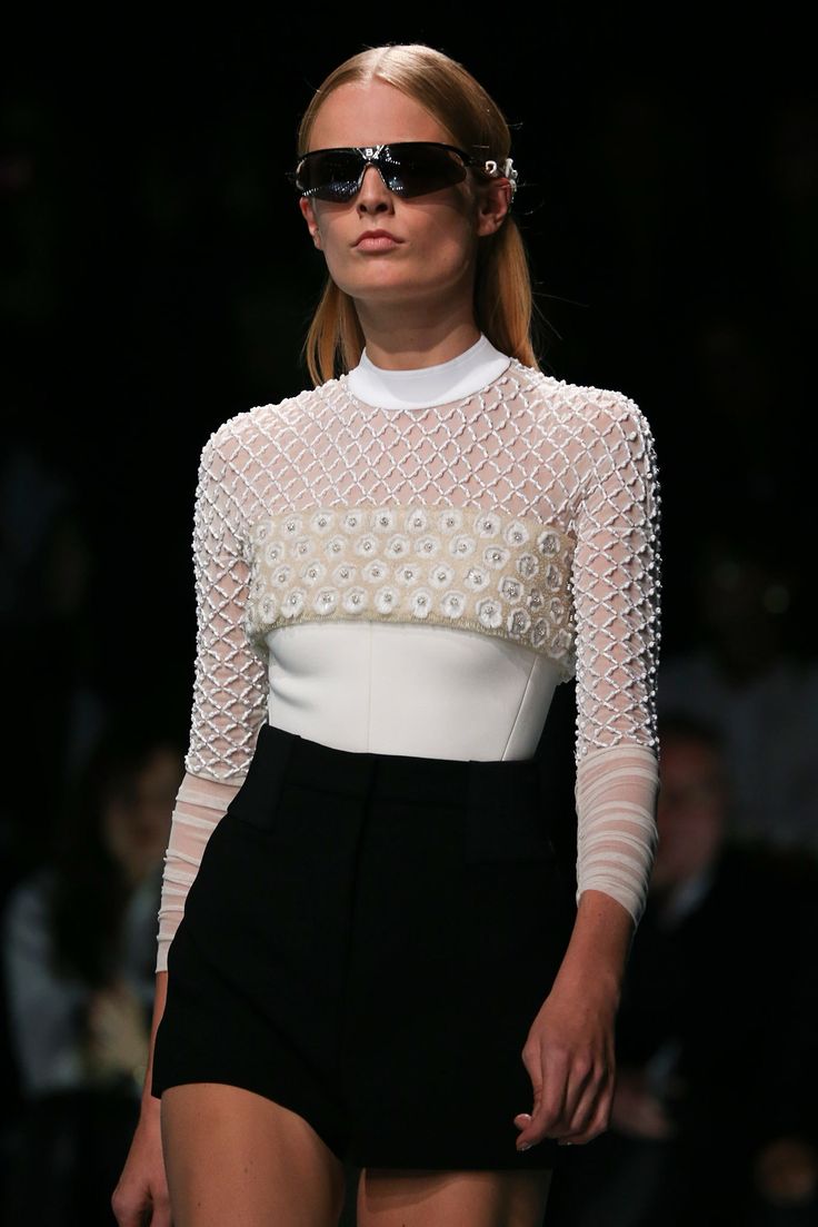 a woman is wearing a white top with a black skirt.  she has on sunglasses.  she is on a runway.