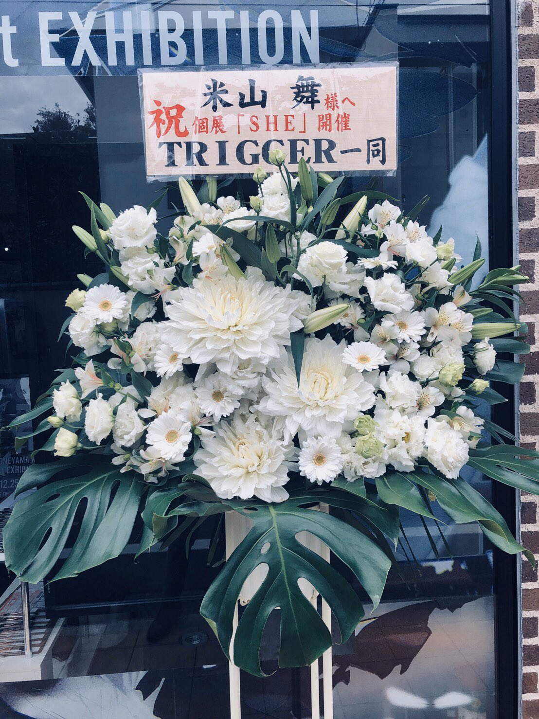 a large floral arrangement of white flowers and lush greenery stands in front of a storefront, congratulating an artist on their exhibition. the message on the sign is written in japanese and expresses congratulations to 米山舞 (yamaguchi mai) from the group trigger for the opening of their solo exhibition titled "she". the storefront suggests an art exhibition space.