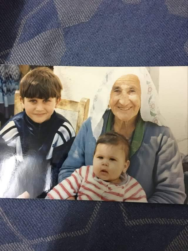 an old, smiling woman is sitting in the middle of the photo. a boy and a baby are sitting on either side of her. the woman has a white scarf on her head, and the baby is wearing a red and white striped shirt. the boy is wearing a black jacket with white stripes on the sleeves. the background is a blurry white wall, and the photo appears to be from the 1990s. the overall feeling of the picture is one of love and family.
