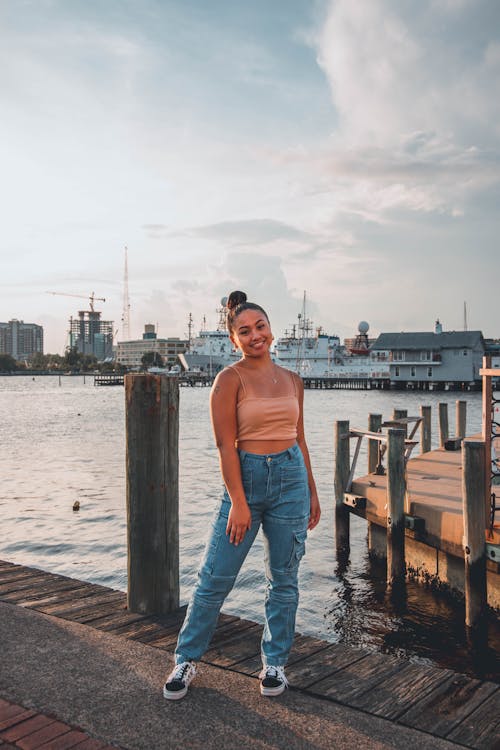 a young woman smiles and poses for a photo on a wooden dock at a harbor. she's wearing a peachcolored tank top and light blue cargo jeans. behind her is a cityscape with buildings, cranes, and several boats and ships in the water. the sky is partly cloudy and there's sunlight reflecting on the water.