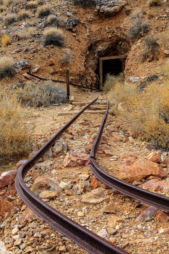 the image shows an abandoned mine shaft in a desert landscape. the entrance to the mine is visible, supported by a wooden frame, and rusty railroad tracks lead into the tunnel. the ground is rocky and covered with gravel, with dry shrubs dotting the arid landscape. the scene suggests a hot, dry environment and evokes a sense of history and abandonment.